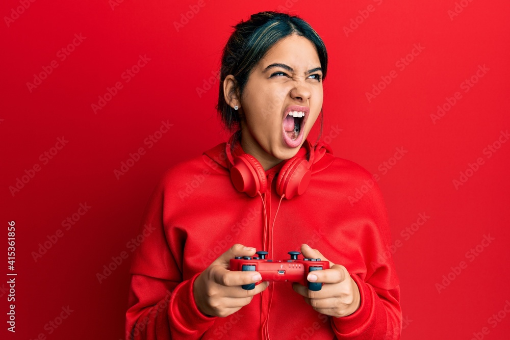 Young latin woman playing video game holding controller angry and mad ...
