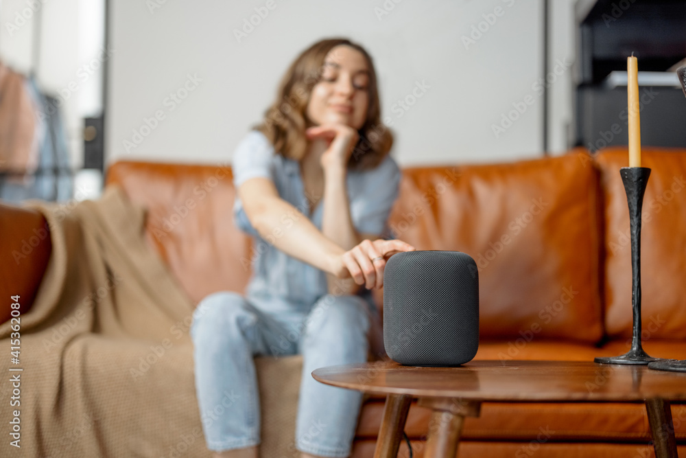 Pretty woman touching black audio assistant column on the coffee table while sitting on foxy leather sofa in stylish interior design of living room. Smart home concept. Pretty woman touching black audio assistant column on the coffee table while sitting on foxy leather sofa in stylish interior design of living room. Smart home concept.