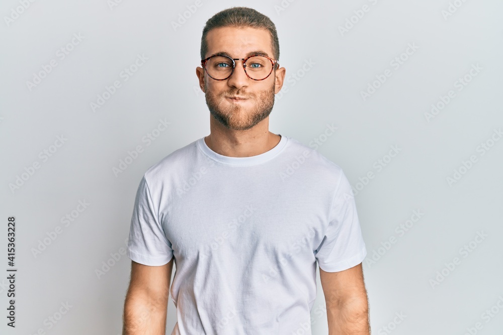 Young caucasian man wearing casual clothes and glasses puffing cheeks with funny face. mouth inflated with air, crazy expression. Young caucasian man wearing casual clothes and glasses puffing cheeks with funny face. mouth inflated with air, crazy expression.