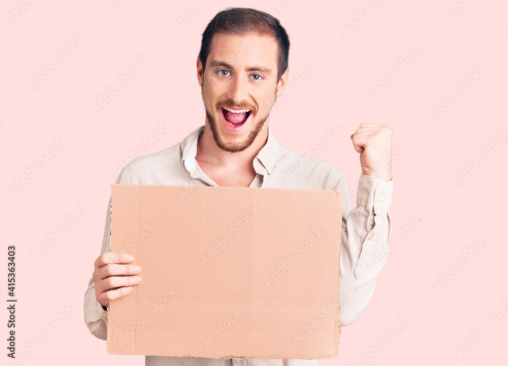 Young handsome caucasian man holding empty cardboard banner screaming proud, celebrating victory and success very excited with raised arms Young handsome caucasian man holding empty cardboard banner screaming proud, celebrating victory and success very excited with raised arms