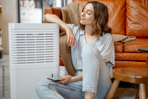 Pretty woman sitting near air purifier and moisturizer appliance near sofa monitoring air quality in phone. Health microclimate at home concept.  photo