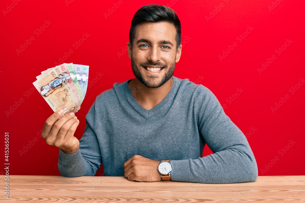Handsome hispanic man holding canadian dollars looking positive and happy standing and smiling with a confident smile showing teeth Handsome hispanic man holding canadian dollars looking positive and happy standing and smiling with a confident smile showing teeth