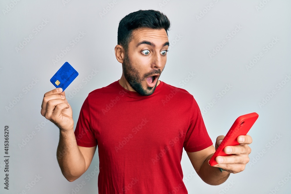 Young hispanic man holding smartphone and credit card afraid and shocked with surprise and amazed expression, fear and excited face. Young hispanic man holding smartphone and credit card afraid and shocked with surprise and amazed expression, fear and excited face.