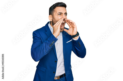 Young hispanic man wearing business jacket shouting angry out loud with hands over mouth