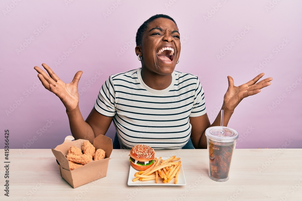 Young african american woman eating a tasty classic burger with fries and soda crazy and mad shouting and yelling with aggressive expression and arms raised. frustration concept. Young african american woman eating a tasty classic burger with fries and soda crazy and mad shouting and yelling with aggressive expression and arms raised. frustration concept.