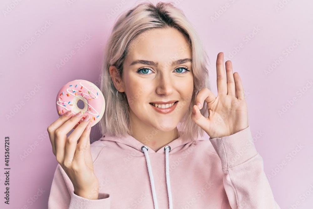 Young blonde girl holding donut doing ok sign with fingers, smiling friendly gesturing excellent symbol Young blonde girl holding donut doing ok sign with fingers, smiling friendly gesturing excellent symbol