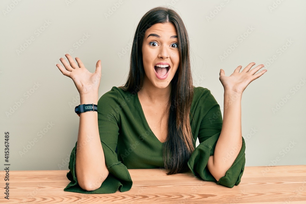 Young brunette woman wearing casual clothes sitting on the table celebrating crazy and amazed for success with arms raised and open eyes screaming excited. winner concept Young brunette woman wearing casual clothes sitting on the table celebrating crazy and amazed for success with arms raised and open eyes screaming excited. winner concept