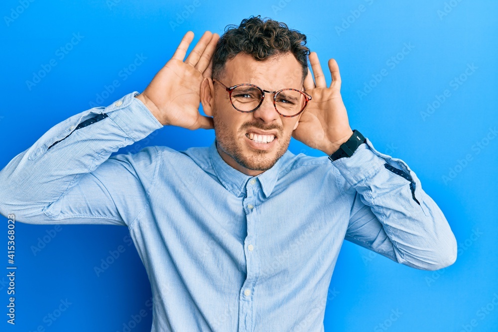 Young hispanic man wearing casual clothes and glasses trying to hear both hands on ear gesture, curious for gossip. hearing problem, deaf Young hispanic man wearing casual clothes and glasses trying to hear both hands on ear gesture, curious for gossip. hearing problem, deaf