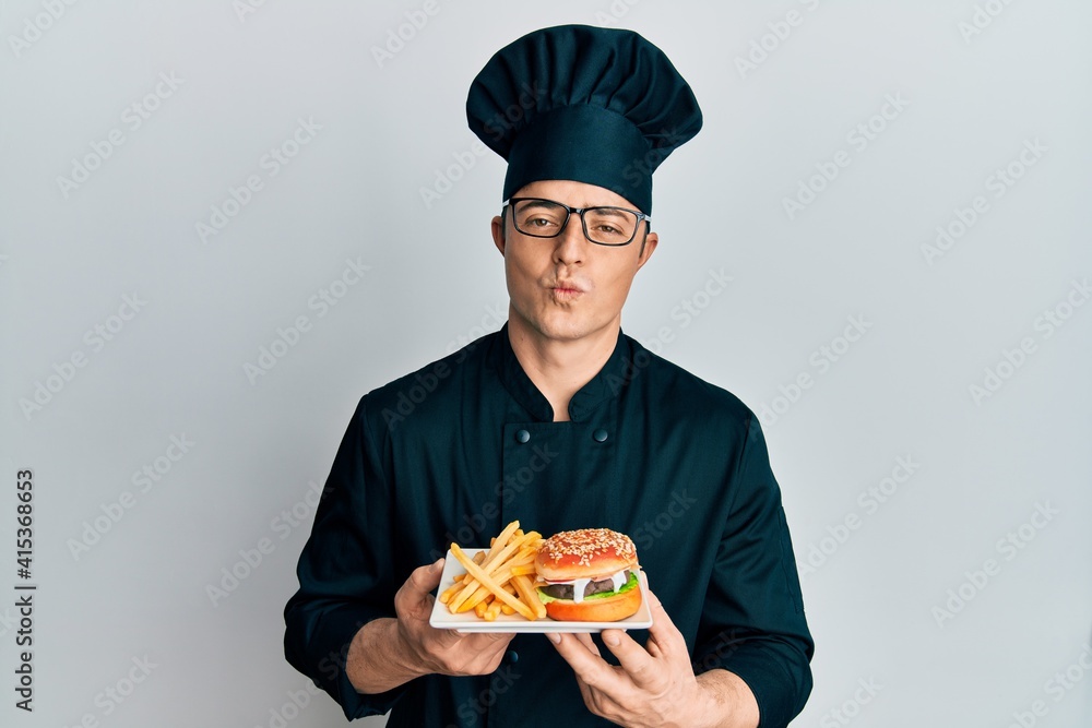 Handsome young man chef holding burger with fries looking at the camera blowing a kiss being lovely and sexy. love expression. Handsome young man chef holding burger with fries looking at the camera blowing a kiss being lovely and sexy. love expression.