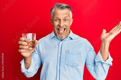 Middle age grey-haired man drinking glass of whisky celebrating victory with happy smile and winner expression with raised hands photo