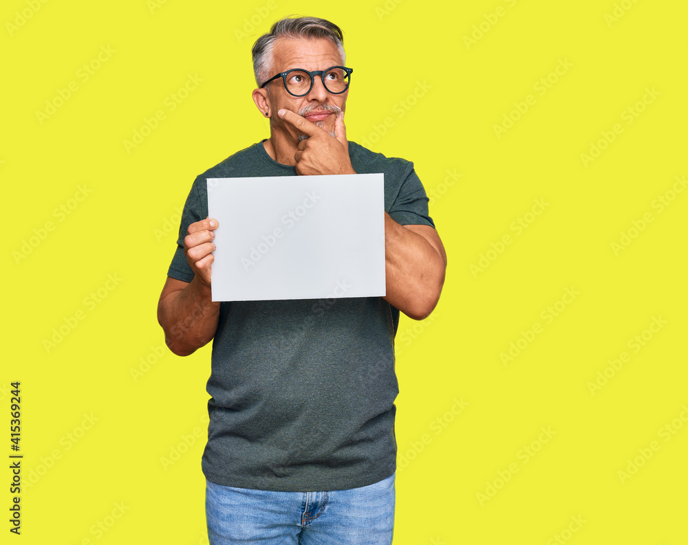 Middle age grey-haired man holding blank empty banner serious face thinking about question with hand on chin, thoughtful about confusing idea Middle age grey-haired man holding blank empty banner serious face thinking about question with hand on chin, thoughtful about confusing idea