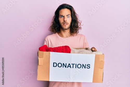 Young handsome man with long hair holding donations box for charity in shock face, looking skeptical and sarcastic, surprised with open mouth © Krakenimages.com