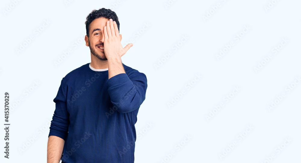 Young hispanic man wearing casual clothes covering one eye with hand, confident smile on face and surprise emotion. Young hispanic man wearing casual clothes covering one eye with hand, confident smile on face and surprise emotion.