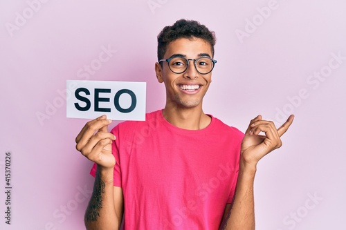 Young handsome african american man holding seo message paper smiling happy pointing with hand and finger to the side © Krakenimages.com