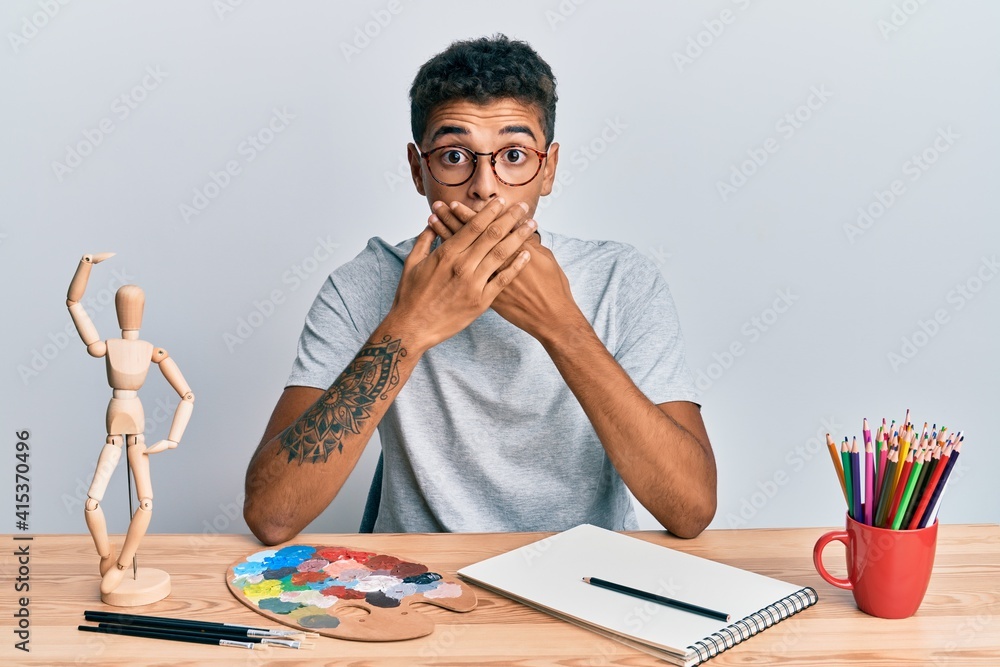 Young handsome african american man painter sitting palette and art manikin shocked covering mouth with hands for mistake. secret concept. Young handsome african american man painter sitting palette and art manikin shocked covering mouth with hands for mistake. secret concept.