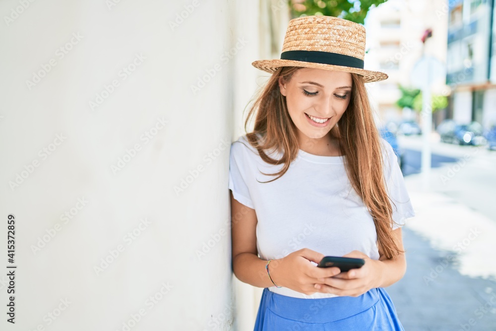 Young blonde woman on vacation smiling happy using smartphone leaning on the wall at street of city