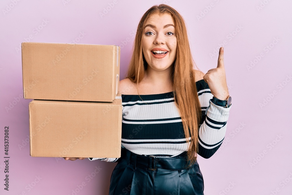 Young beautiful redhead woman holding delivery package smiling with an idea or question pointing finger with happy face, number one Young beautiful redhead woman holding delivery package smiling with an idea or question pointing finger with happy face, number one