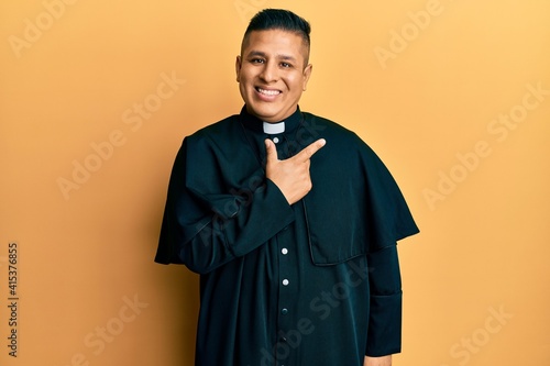 Young latin priest man standing over yellow background cheerful with a smile on face pointing with hand and finger up to the side with happy and natural expression