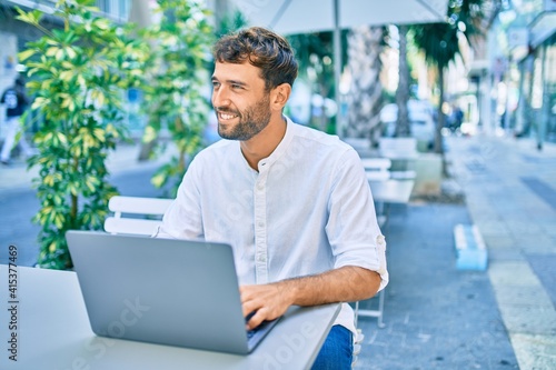 Handsome man with beard wearing casual white shirt on a sunny day working using laptop at cafeteria