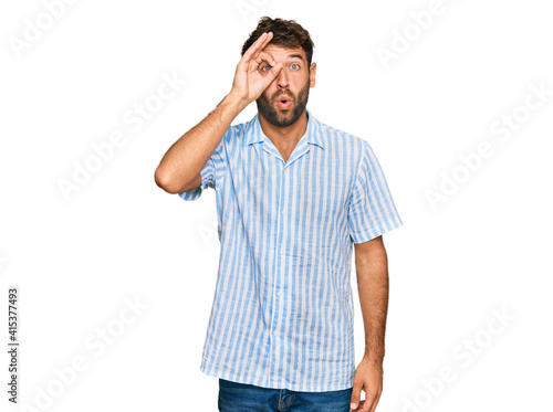 Handsome young man with beard wearing casual fresh shirt doing ok gesture shocked with surprised face, eye looking through fingers. unbelieving expression.