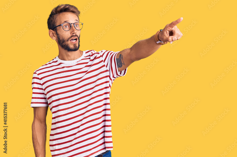Fototapeta premium Handsome blond man with beard wearing casual clothes and glasses pointing with finger surprised ahead, open mouth amazed expression, something on the front