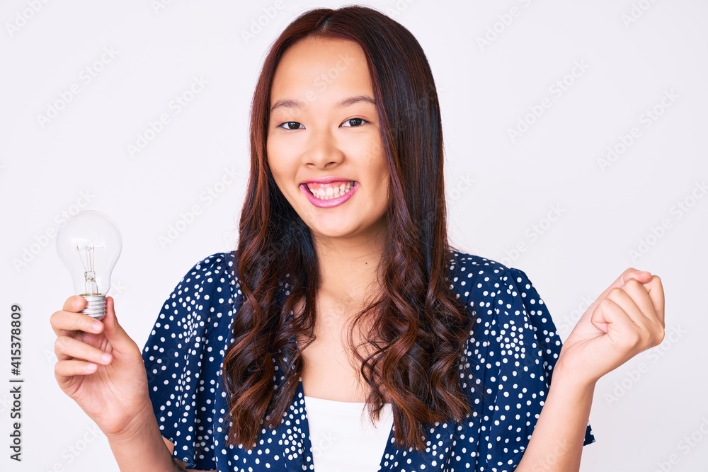 Young beautiful chinese girl holding lightbulb for inspiration and idea screaming proud, celebrating victory and success very excited with raised arm Young beautiful chinese girl holding lightbulb for inspiration and idea screaming proud, celebrating victory and success very excited with raised arm