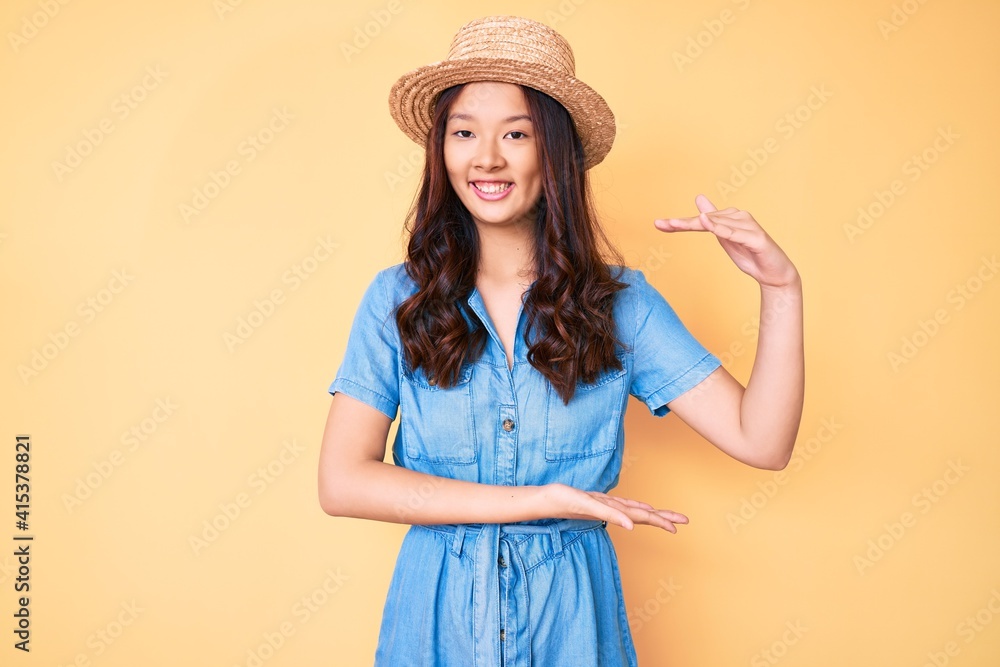 Young beautiful chinese girl wearing summer hat gesturing with hands showing big and large size sign, measure symbol. smiling looking at the camera. measuring concept. Young beautiful chinese girl wearing summer hat gesturing with hands showing big and large size sign, measure symbol. smiling looking at the camera. measuring concept.