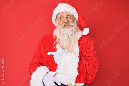 Old senior man with grey hair and long beard wearing traditional santa claus costume cheerful with a smile of face pointing with hand and finger up to the side with happy and natural expression
