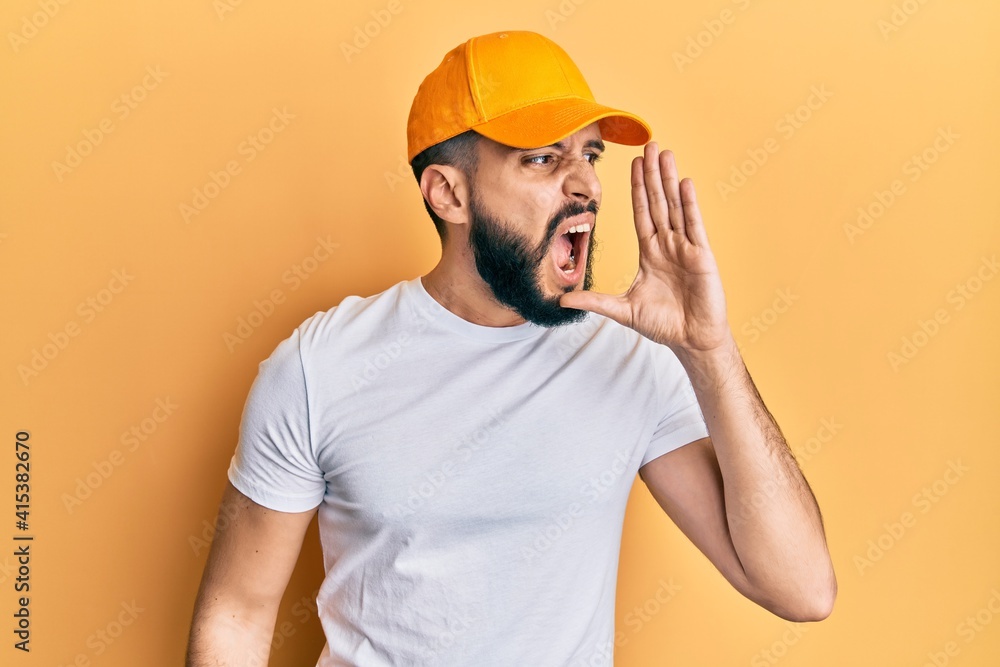 Young man with beard wearing yellow cap shouting and screaming loud to side with hand on mouth. communication concept.