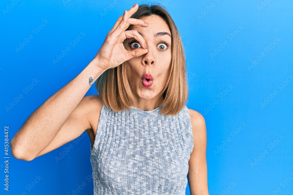 Young caucasian blonde woman wearing casual t shirt doing ok gesture shocked with surprised face, eye looking through fingers. unbelieving expression.