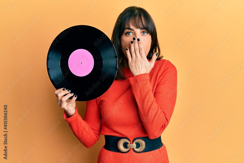 Fototapeta premium Young hispanic woman holding vinyl disc covering mouth with hand, shocked and afraid for mistake. surprised expression