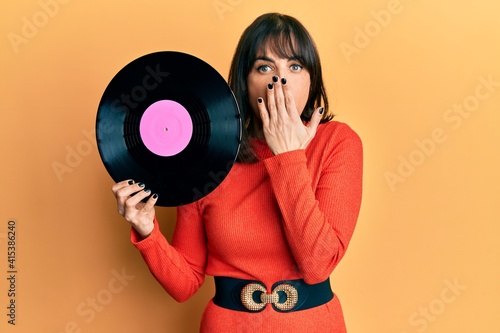 Young hispanic woman holding vinyl disc covering mouth with hand, shocked and afraid for mistake. surprised expression photo