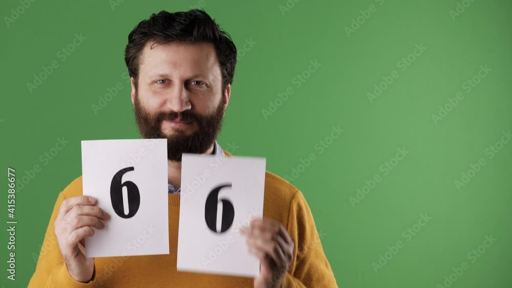 Man holding number 66 sign. Cheerful white bearded good-looking man in yellow sweater shows number sixty-six on white sheet. Chrome key, green screen, copy space Man holding number 66 sign. Cheerful white bearded good-looking man in yellow sweater shows number sixty-six on white sheet. Chrome key, green screen, copy space