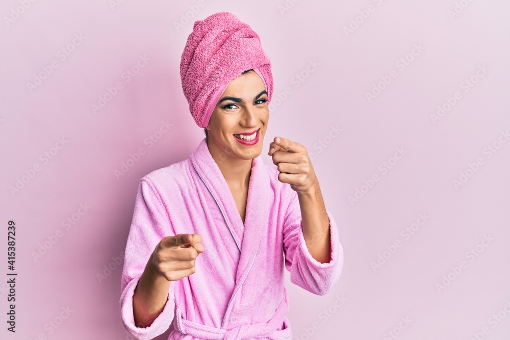 Young man wearing woman make up wearing shower towel on head and bathrobe pointing fingers to camera with happy and funny face. good energy and vibes. Young man wearing woman make up wearing shower towel on head and bathrobe pointing fingers to camera with happy and funny face. good energy and vibes.