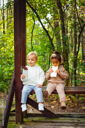 A little blonde boy and an African-American girl in a park on a bench eating a bar of white and dark chocolate. © Elena 