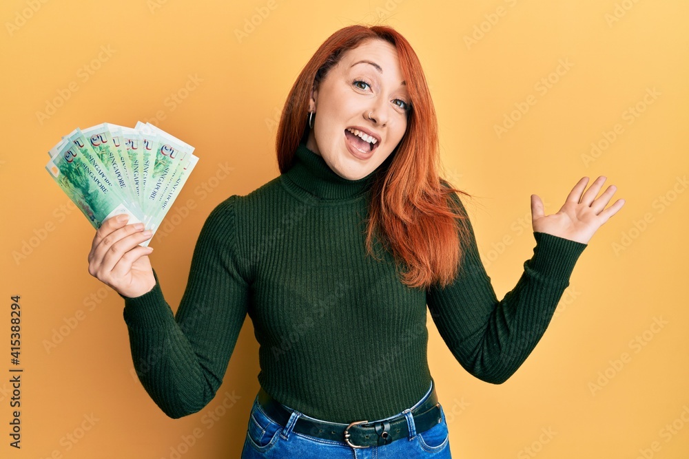 Beautiful redhead woman holding 5 singapore dollars banknotes celebrating achievement with happy smile and winner expression with raised hand Beautiful redhead woman holding 5 singapore dollars banknotes celebrating achievement with happy smile and winner expression with raised hand