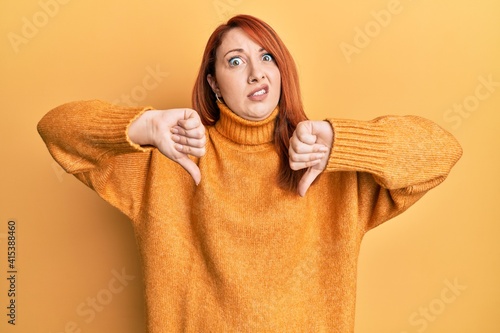 Quadro su tela Beautiful redhead woman doing negative thumbs down gesture in shock face, lookin