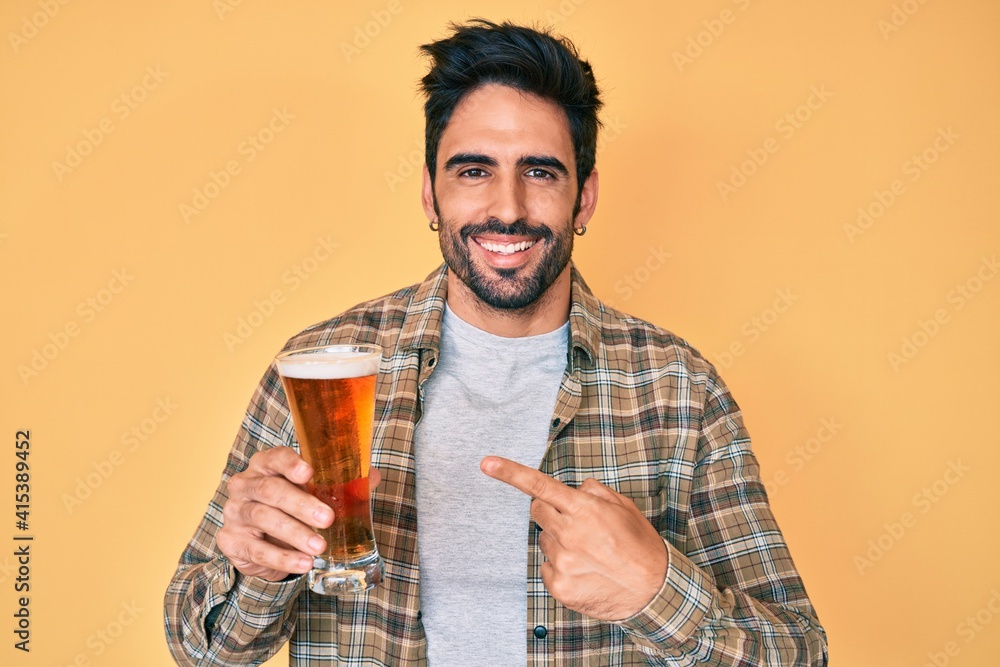 Handsome hispanic man with beard drinking a pint of beer smiling happy pointing with hand and finger Handsome hispanic man with beard drinking a pint of beer smiling happy pointing with hand and finger
