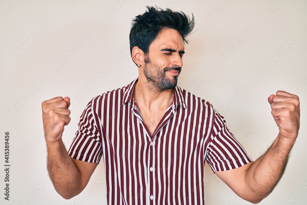 Fototapeta premium Handsome hispanic man with beard wearing casual clothes very happy and excited doing winner gesture with arms raised, smiling and screaming for success. celebration concept.