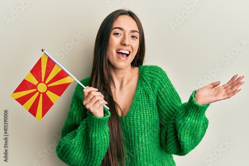 Young hispanic girl holding macedonian flag celebrating achievement with happy smile and winner expression with raised hand