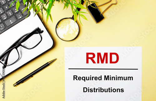 RMD Required Minimum Distributions is written in red on a white piece of paper on a light yellow background next to a laptop, pen, magnifying glass, glasses and a green plant. photo