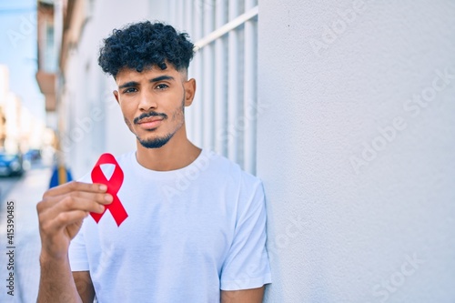 Young arab man with serious expression holding hiv awaraness red ribbon leaning on the wall. photo