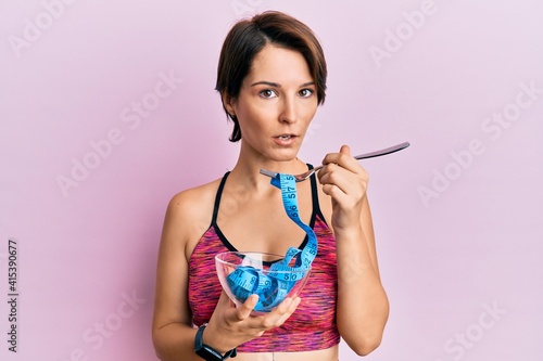Young brunette woman with short hair eating measure meter doing diet for weight loss clueless and confused expression. doubt concept.