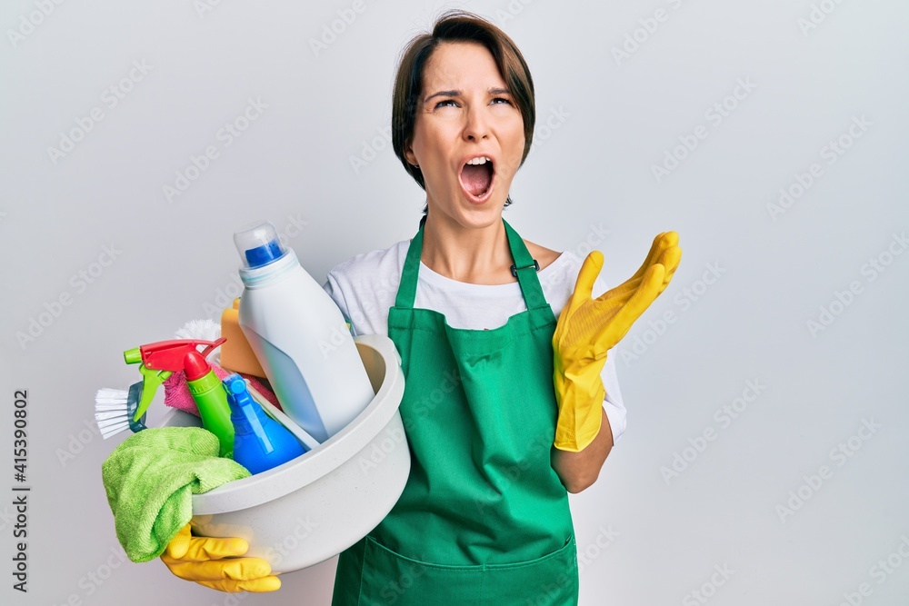 Young brunette woman with short hair wearing apron holding cleaning products crazy and mad shouting and yelling with aggressive expression and arms raised. frustration concept. Young brunette woman with short hair wearing apron holding cleaning products crazy and mad shouting and yelling with aggressive expression and arms raised. frustration concept.