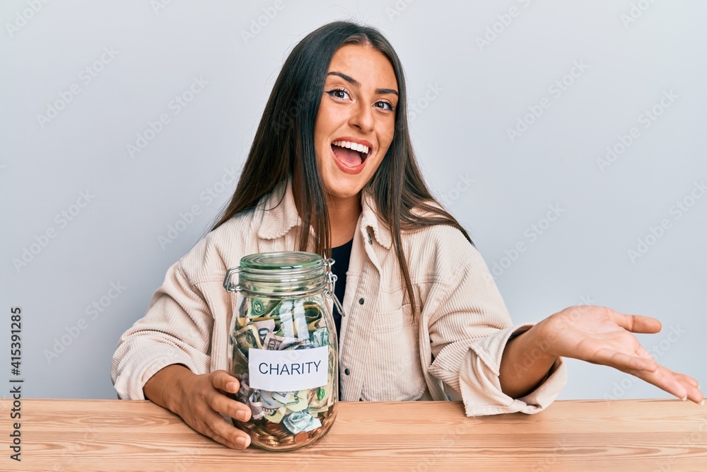 Beautiful hispanic woman holding charity jar with money celebrating achievement with happy smile and winner expression with raised hand Beautiful hispanic woman holding charity jar with money celebrating achievement with happy smile and winner expression with raised hand