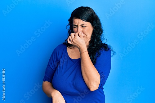 Plus size brunette woman wearing casual blue shirt smelling something stinky and disgusting, intolerable smell, holding breath with fingers on nose. bad smell