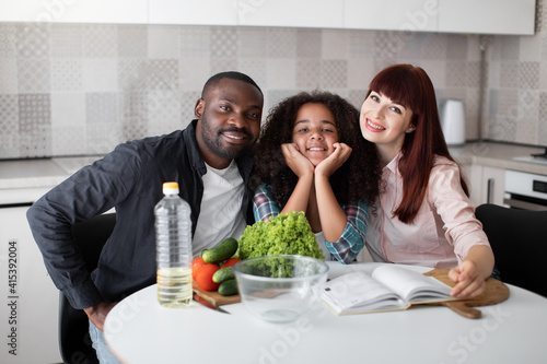 Happy multiethnic family with teen mixed raced daughter  looking at the camera with pleasure smiles  sitting at the table in kitchen interior