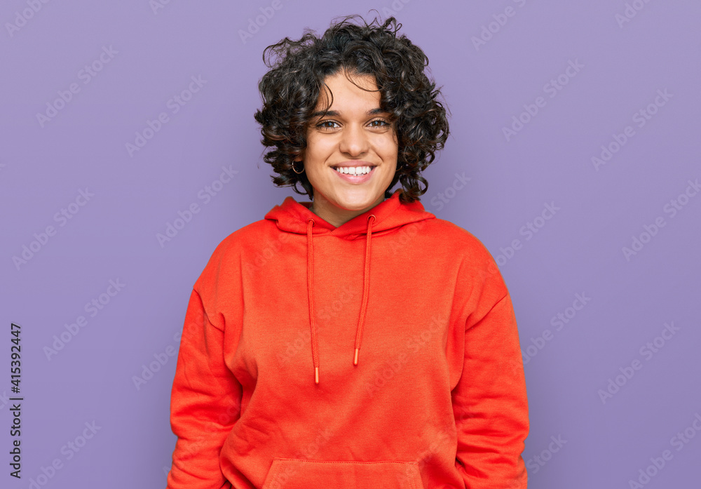 Young hispanic woman with curly hair wearing casual sweatshirt looking positive and happy standing and smiling with a confident smile showing teeth Young hispanic woman with curly hair wearing casual sweatshirt looking positive and happy standing and smiling with a confident smile showing teeth
