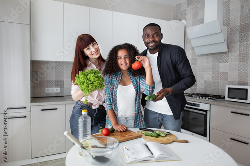Cheerful young pleasant multiethnic family with cute daughter, showing fresh vegetables, cucumber, tomato and lettuse to the camera, posing at home kitchen photo