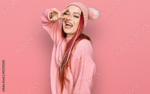 Young caucasian woman wearing casual clothes and wool cap doing peace symbol with fingers over face, smiling cheerful showing victory